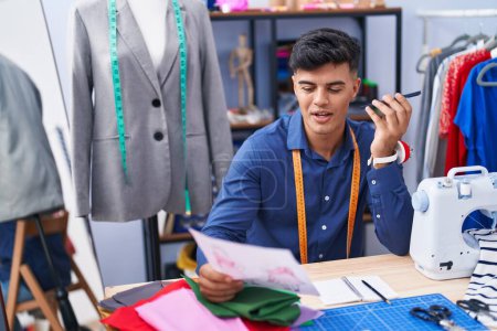 Téléchargez les photos : Jeune homme hispanique tailleur parler sur smartphone regarder conception de vêtements à l'usine de vêtements - en image libre de droit