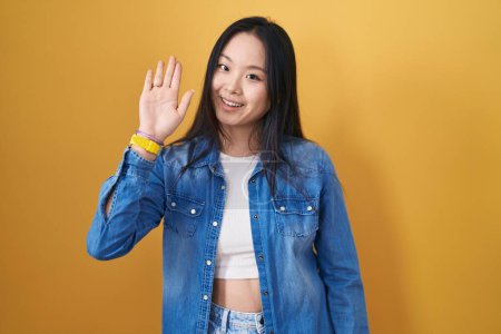 Téléchargez les photos : Jeune femme asiatique debout sur fond jaune renonçant à dire bonjour heureux et souriant, geste de bienvenue amical - en image libre de droit