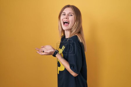 Foto de Mujer rubia caucásica usando camiseta bitcoin señalando a un lado con las manos abiertas palmeras mostrando espacio de copia, presentando anuncio sonriendo emocionado feliz - Imagen libre de derechos