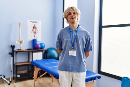 Téléchargez les photos : Jeune homme blond pysiothérapeute souriant confiant debout à la clinique de réadaptation - en image libre de droit