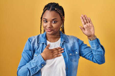 Foto de Mujer afroamericana con trenzas de pie sobre fondo amarillo jurando con la mano en el pecho y la palma abierta, haciendo un juramento de lealtad promesa - Imagen libre de derechos