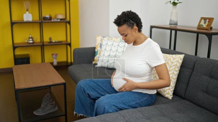 Téléchargez les photos : Jeune femme enceinte touchant le ventre assis sur un canapé avec une expression triste à la maison - en image libre de droit