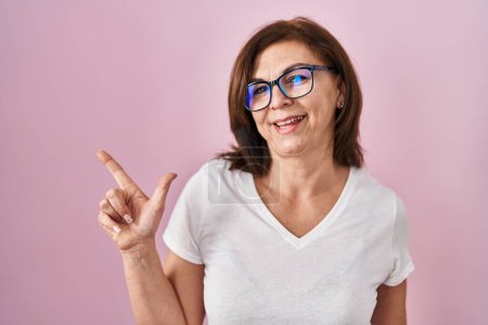 Foto de Mujer hispana de mediana edad de pie sobre fondo rosa con una gran sonrisa en la cara, señalando con el dedo de la mano hacia el lado mirando a la cámara. - Imagen libre de derechos