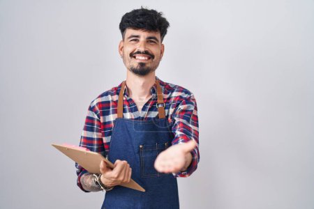Photo for Young hispanic man with beard wearing waiter apron holding clipboard smiling cheerful offering palm hand giving assistance and acceptance. - Royalty Free Image