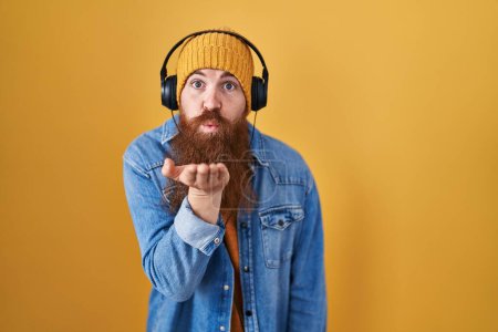 Foto de Hombre caucásico con barba larga escuchando música usando auriculares mirando a la cámara soplando un beso con la mano en el aire siendo encantador y sexy. expresión de amor. - Imagen libre de derechos