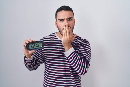 Photo for Young hispanic man wearing pyjama holding alarm clock covering mouth with hand, shocked and afraid for mistake. surprised expression - Royalty Free Image