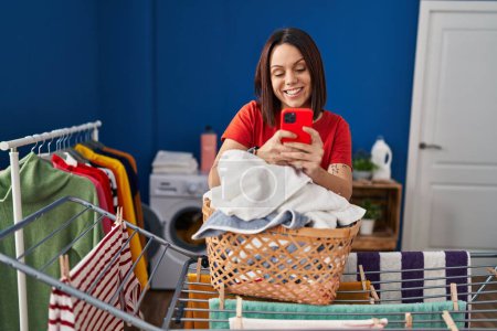 Photo for Young beautiful hispanic woman using smartphone hanging clothes on clothesline at laundry room - Royalty Free Image