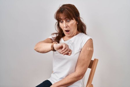 Téléchargez les photos : Femme hispanique du Moyen Âge se faire vacciner montrant bras avec pansement regardant l'heure de la montre inquiet, peur de se faire tard - en image libre de droit