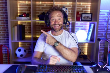 Photo for Middle age man with beard playing video games wearing headphones cheerful with a smile on face pointing with hand and finger up to the side with happy and natural expression - Royalty Free Image