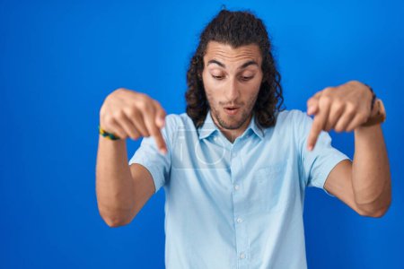 Photo for Young hispanic man standing over blue background pointing down with fingers showing advertisement, surprised face and open mouth - Royalty Free Image