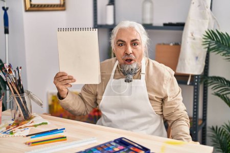 Foto de Hombre de mediana edad con pelo gris sentado en el estudio de arte sosteniendo portátil asustado y sorprendido con la boca abierta para sorpresa, cara de incredulidad - Imagen libre de derechos
