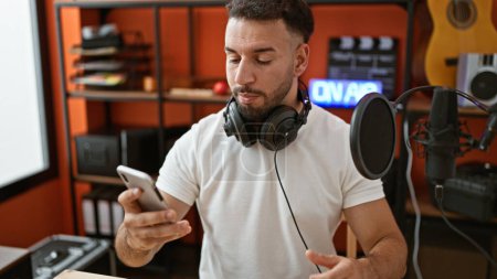 Téléchargez les photos : Jeune homme arabe musicien portant un casque en utilisant un smartphone au studio de musique - en image libre de droit