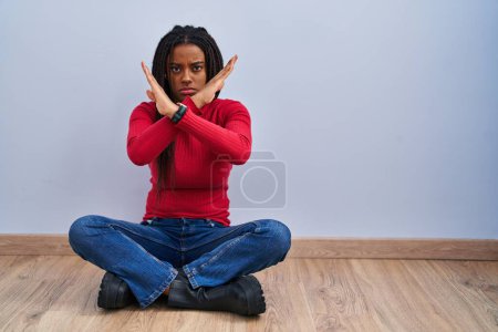 Photo for Young african american with braids sitting on the floor at home rejection expression crossing arms doing negative sign, angry face - Royalty Free Image