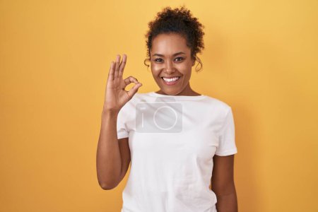 Photo for Young hispanic woman with curly hair standing over yellow background smiling positive doing ok sign with hand and fingers. successful expression. - Royalty Free Image