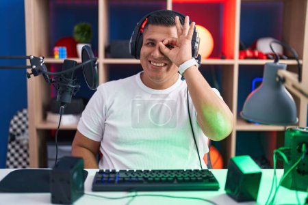 Foto de Joven hispano jugando videojuegos haciendo buen gesto con la mano sonriente, los ojos mirando a través de los dedos con la cara feliz. - Imagen libre de derechos