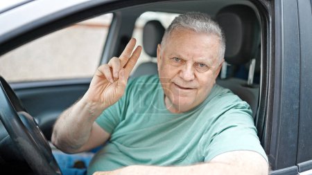 Téléchargez les photos : Moyen Age homme aux cheveux gris souriant confiant assis sur la voiture faisant geste de victoire dans la rue - en image libre de droit