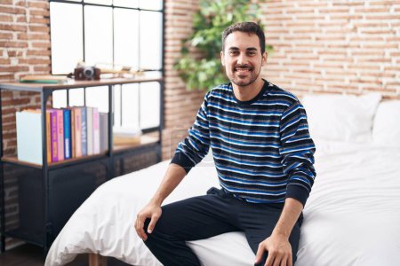 Foto de Young hispanic man smiling confident sitting on bed at bedroom - Imagen libre de derechos