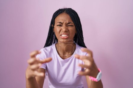 Téléchargez les photos : Femme afro-américaine avec des tresses debout sur fond rose criant frustré par la rage, les mains essayant d'étrangler, hurlant fou - en image libre de droit