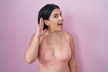 Téléchargez les photos : Jeune femme hispanique portant un soutien-gorge rose souriant avec la main sur l'oreille en écoutant une rumeur ou des commérages. concept de surdité. - en image libre de droit