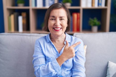 Photo for Middle age hispanic woman sitting on the sofa at home cheerful with a smile of face pointing with hand and finger up to the side with happy and natural expression on face - Royalty Free Image