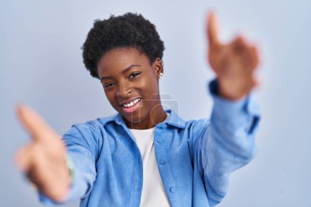 Téléchargez les photos : Femme afro-américaine debout sur fond bleu regardant la caméra souriant à bras ouverts pour un câlin. expression joyeuse embrassant le bonheur. - en image libre de droit