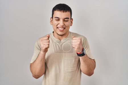 Téléchargez les photos : Jeune homme arabe portant t-shirt décontracté excité pour le succès avec les bras levés et les yeux fermés célébrant la victoire en souriant. concept gagnant. - en image libre de droit