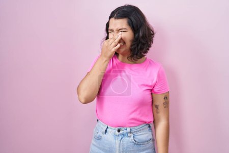 Téléchargez les photos : Jeune femme hispanique debout sur fond rose sentant quelque chose de puant et dégoûtant, odeur intolérable, retenant souffle avec les doigts sur le nez. mauvaise odeur - en image libre de droit