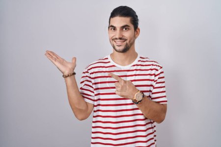 Photo for Hispanic man with long hair standing over isolated background amazed and smiling to the camera while presenting with hand and pointing with finger. - Royalty Free Image