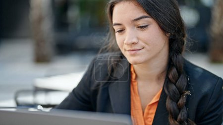 Téléchargez les photos : Jeune belle ouvrière hispanique utilisant un ordinateur portable avec une expression détendue sur la terrasse du café - en image libre de droit