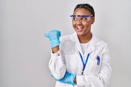 Foto de Mujer afroamericana con trenzas vistiendo bata científica sonriendo con la cara feliz mirando y señalando hacia un lado con el pulgar hacia arriba. - Imagen libre de derechos
