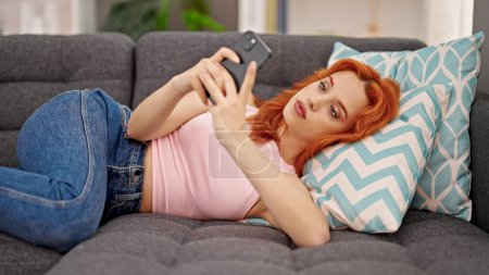 Foto de Young redhead woman using smartphone lying on sofa at home - Imagen libre de derechos