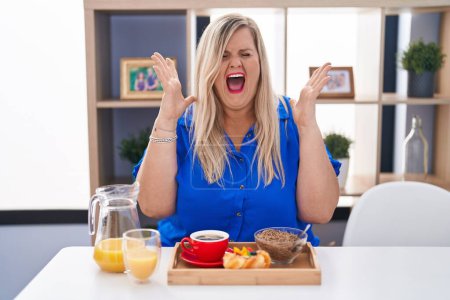 Foto de Mujer caucásica de talla grande desayunando en casa celebrando locos y locos por el éxito con los brazos levantados y los ojos cerrados gritando emocionados. concepto ganador - Imagen libre de derechos