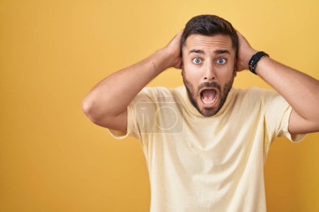 Handsome hispanic man standing over yellow background crazy and scared with hands on head, afraid and surprised of shock with open mouth 