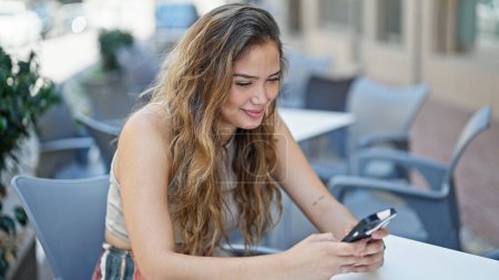 Foto de Joven mujer hispana hermosa usando teléfono inteligente sentado en la mesa sonriendo en la terraza de la cafetería - Imagen libre de derechos