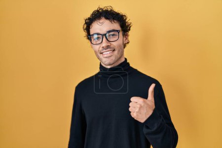 Téléchargez les photos : Homme hispanique debout sur fond jaune faisant des pouces heureux geste avec la main. approuvant l'expression regardant la caméra montrant le succès. - en image libre de droit