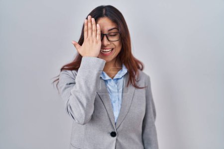 Téléchargez les photos : Jeune femme d'affaires hispanique portant des lunettes couvrant un oeil avec la main, sourire confiant sur le visage et l'émotion surprise. - en image libre de droit