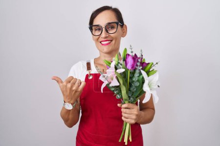 Foto de Mujer morena de mediana edad usando delantal trabajando en floristería sosteniendo ramo apuntando a la espalda con la mano y los pulgares hacia arriba, sonriendo confiado - Imagen libre de derechos