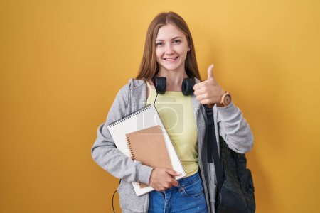 Photo for Young caucasian woman wearing student backpack and holding books doing happy thumbs up gesture with hand. approving expression looking at the camera showing success. - Royalty Free Image