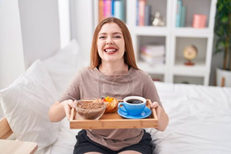 Téléchargez les photos : Femme rousse portant un pyjama tenant un plateau de petit déjeuner souriant et riant fort à haute voix parce que drôle blague folle. - en image libre de droit