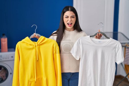 Photo for Young woman hanging clothes on racks angry and mad screaming frustrated and furious, shouting with anger looking up. - Royalty Free Image