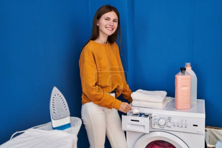 Foto de Young blonde woman pouring detergent on washing machine at laundry room - Imagen libre de derechos