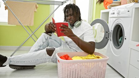 Photo for African american man sitting on floor reading notebook at laundry room - Royalty Free Image