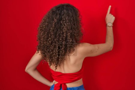 Téléchargez les photos : Femme hispanique avec les cheveux bouclés debout sur fond rouge posant vers l'arrière pointant vers l'avant avec la main du doigt - en image libre de droit