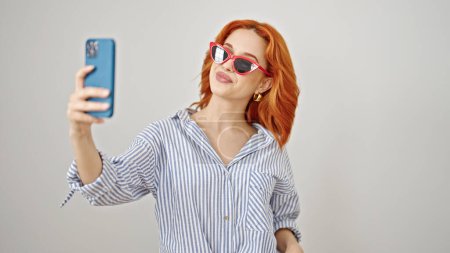 Foto de Mujer pelirroja joven con gafas de sol hacen selfie por teléfono inteligente sobre fondo blanco aislado - Imagen libre de derechos