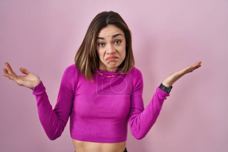 Photo for Hispanic woman standing over pink background clueless and confused expression with arms and hands raised. doubt concept. - Royalty Free Image