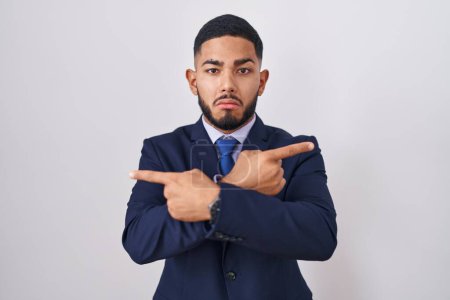 Foto de Joven hombre hispano con traje de negocios y corbata apuntando a ambos lados con los dedos, en diferente dirección en desacuerdo - Imagen libre de derechos