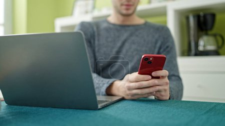 Photo for Young hispanic man using smartphone and laptop sitting on the table at dinning room - Royalty Free Image