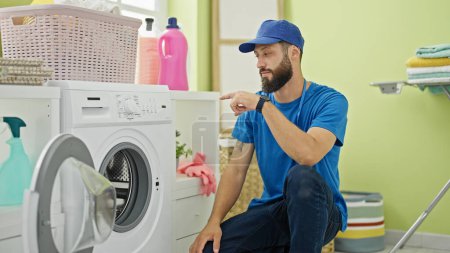 Foto de Joven técnico hispano señalando a la lavadora sonriendo en la lavandería - Imagen libre de derechos