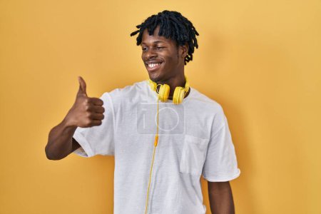Téléchargez les photos : Jeune homme africain avec dreadlocks debout sur fond jaune regardant fier, souriant faire les pouces vers le haut geste sur le côté - en image libre de droit