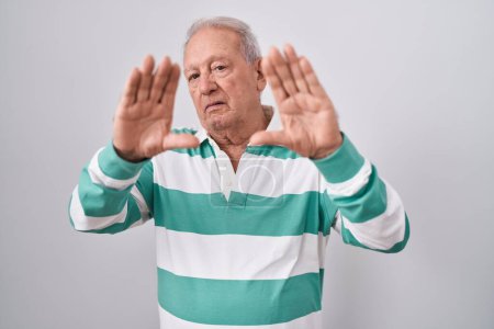 Téléchargez les photos : Homme âgé avec les cheveux gris debout sur fond blanc faisant cadre en utilisant les mains paumes et les doigts, perspective de la caméra - en image libre de droit